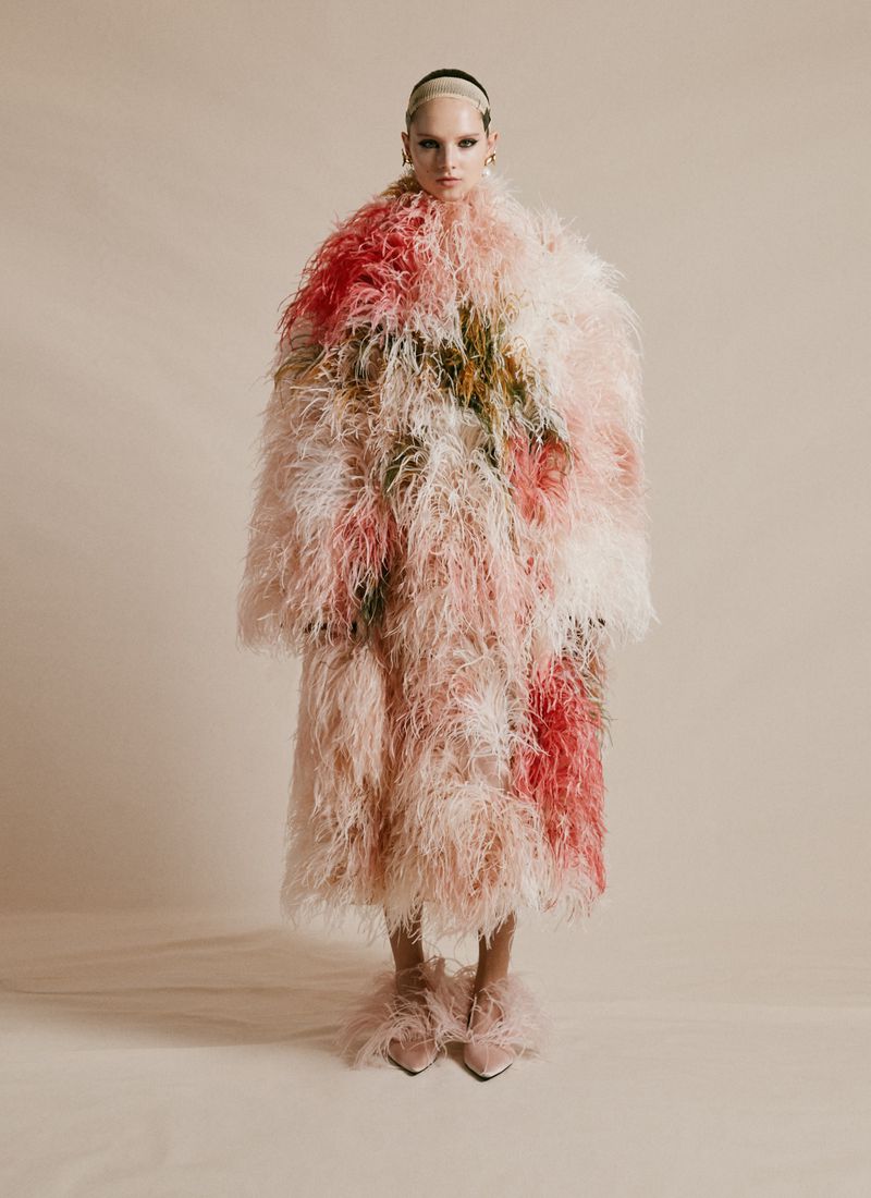 A model in a long pink and white feather coat stands against a neutral backdrop in a fashion shot showcasing unique textures.