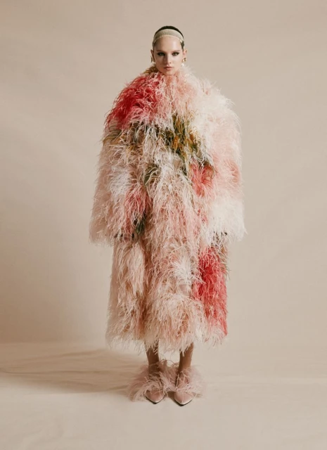 A model in a long pink and white feather coat stands against a neutral backdrop in a fashion shot showcasing unique textures.