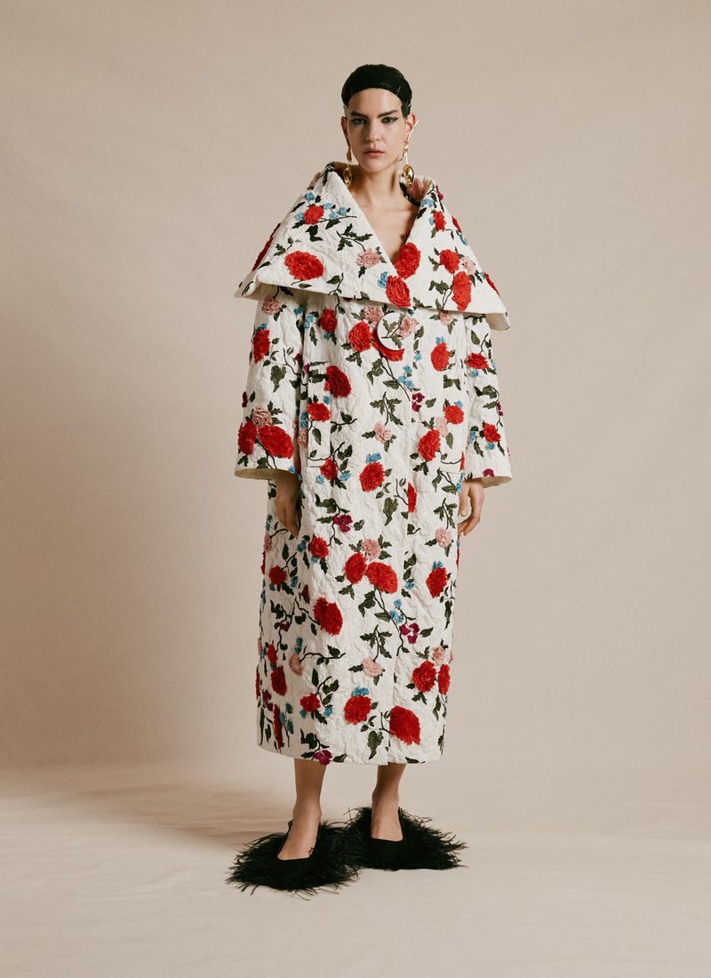 Woman poses in a full-length floral coat with a statement collar, wearing fuzzy slippers against a neutral backdrop in a studio setting.