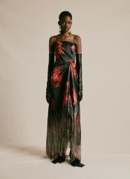 Black model standing in a black and red floral print strapless dress with long fringe hem and black opera gloves against a beige backdrop.