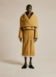 Model standing in a structured mustard yellow wool coat with a large collar and floral brooch against a neutral studio backdrop