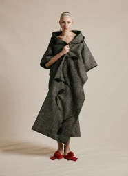 Model posing in an oversized grey tweed cape coat with black floral embellishments and red rosette shoes on a beige studio backdrop.