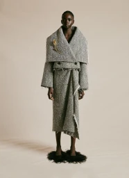 Model wearing a structured tweed coat and matching skirt with feathered black shoes against a neutral background