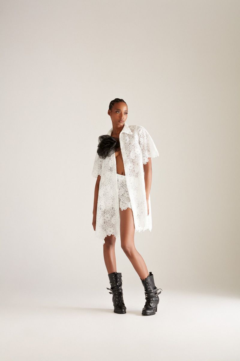 Fashionable adult female poses in a white lace co-ord set with a black floral embellishment and black buckled boots on a plain background.