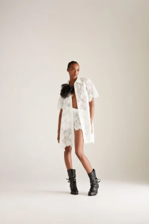 Fashionable adult female poses in a white lace co-ord set with a black floral embellishment and black buckled boots on a plain background.