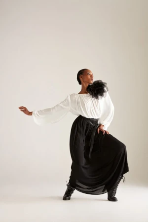 Model Goldie Carswell posing in a white blouse with a black shoulder rosette and long black skirt against a white studio backdrop.