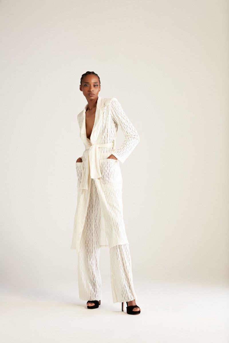 Full-length studio shot of a Black woman in a white lace robe-style outfit with a belt, standing against a neutral background.