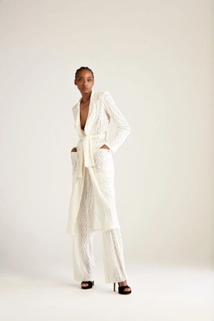 Full-length studio shot of a Black woman in a white lace robe-style outfit with a belt, standing against a neutral background.