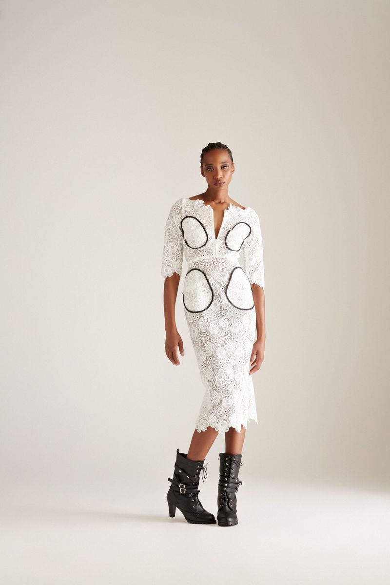 A woman poses in a knee-length white lace dress with black accents and black lace-up heeled boots against a light neutral backdrop.