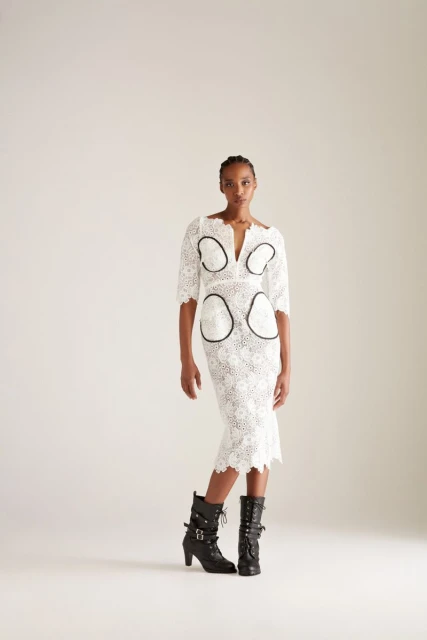 A woman poses in a knee-length white lace dress with black accents and black lace-up heeled boots against a light neutral backdrop.