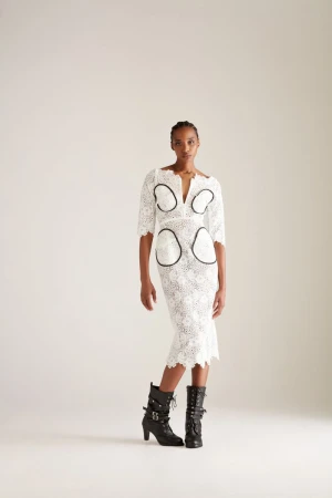 A woman poses in a knee-length white lace dress with black accents and black lace-up heeled boots against a light neutral backdrop.