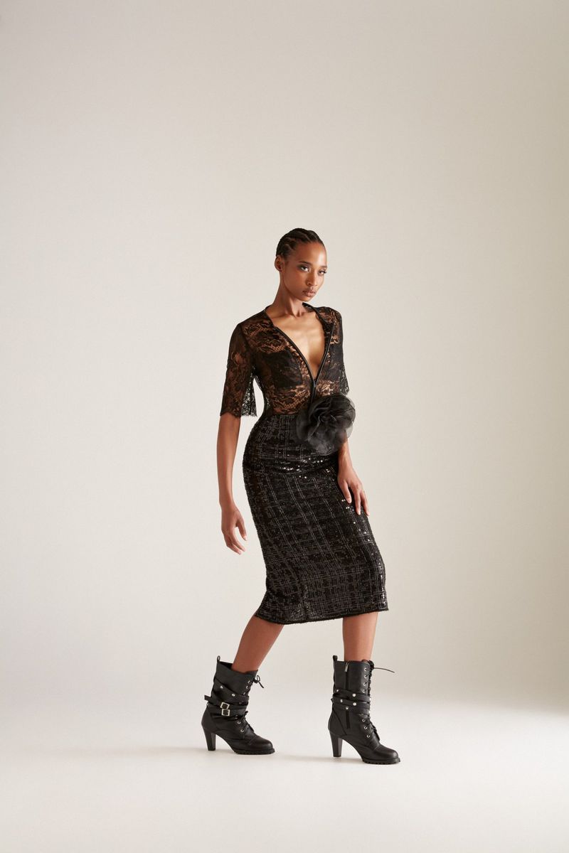 Model Goldie Carswell wearing a black sheer lace midi dress and buckled heeled boots against an off-white seamless studio backdrop.