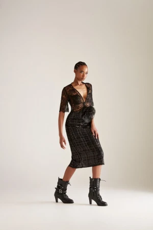 Model Goldie Carswell wearing a black sheer lace midi dress and buckled heeled boots against an off-white seamless studio backdrop.