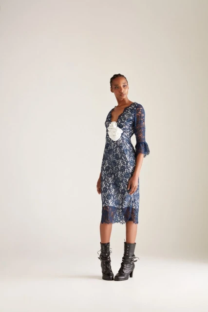 Full-length shot of a Black female fashion model in a blue lace dress with black heeled boots against an off-white studio backdrop.