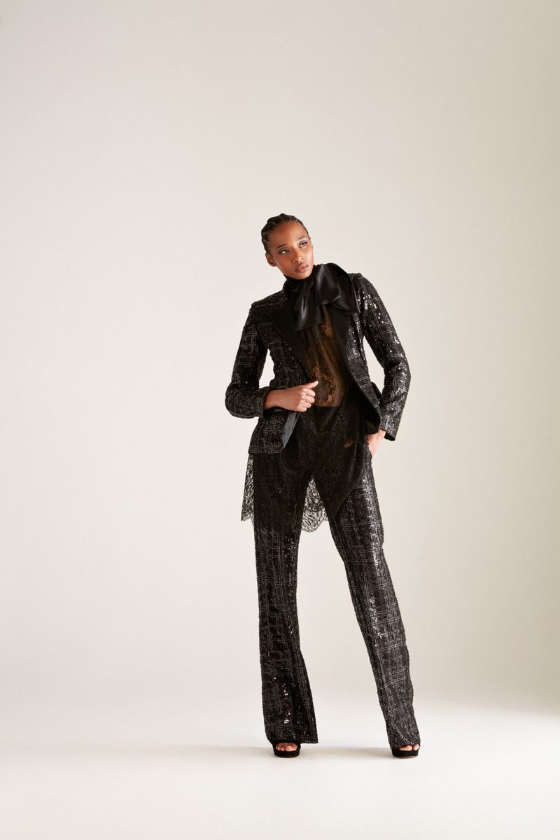 A stylish Black woman stands in a photo studio wearing a dark, sparkling pantsuit, a sheer top, and black platform heels against a white background.