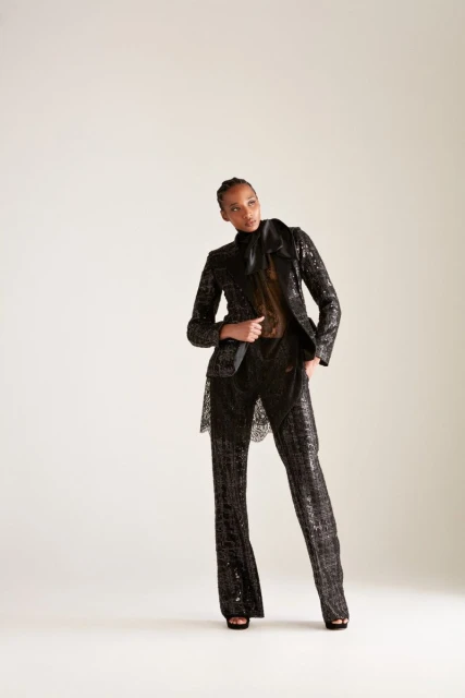 A stylish Black woman stands in a photo studio wearing a dark, sparkling pantsuit, a sheer top, and black platform heels against a white background.