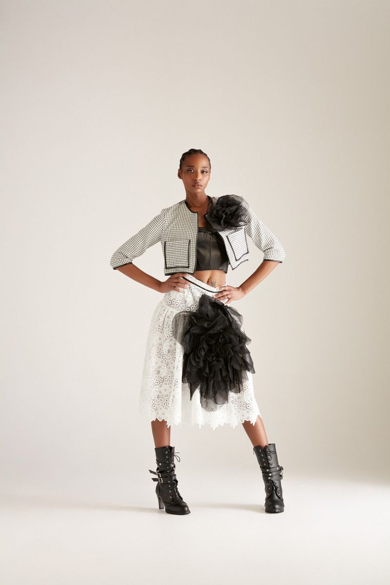 Woman in a fashion pose wearing a striped jacket, crop top, lace skirt with black ruffles, and heeled boots against a white background.