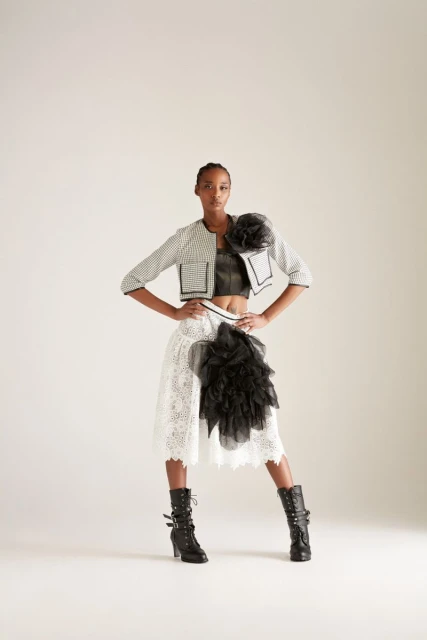 Woman in a fashion pose wearing a striped jacket, crop top, lace skirt with black ruffles, and heeled boots against a white background.
