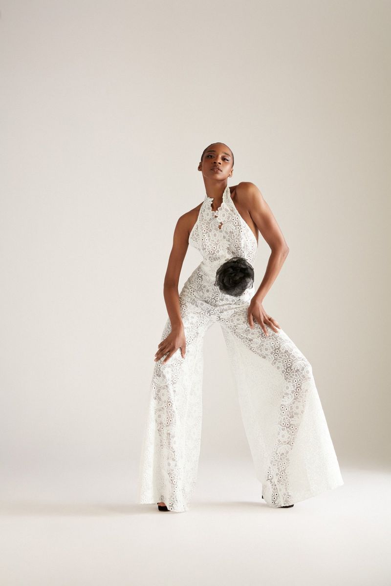 Full-length shot of a slender woman in a white lace halter jumpsuit, posing in a photo studio with hands on knees, looking upward.