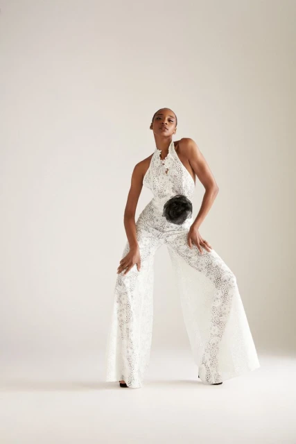 Full-length shot of a slender woman in a white lace halter jumpsuit, posing in a photo studio with hands on knees, looking upward.