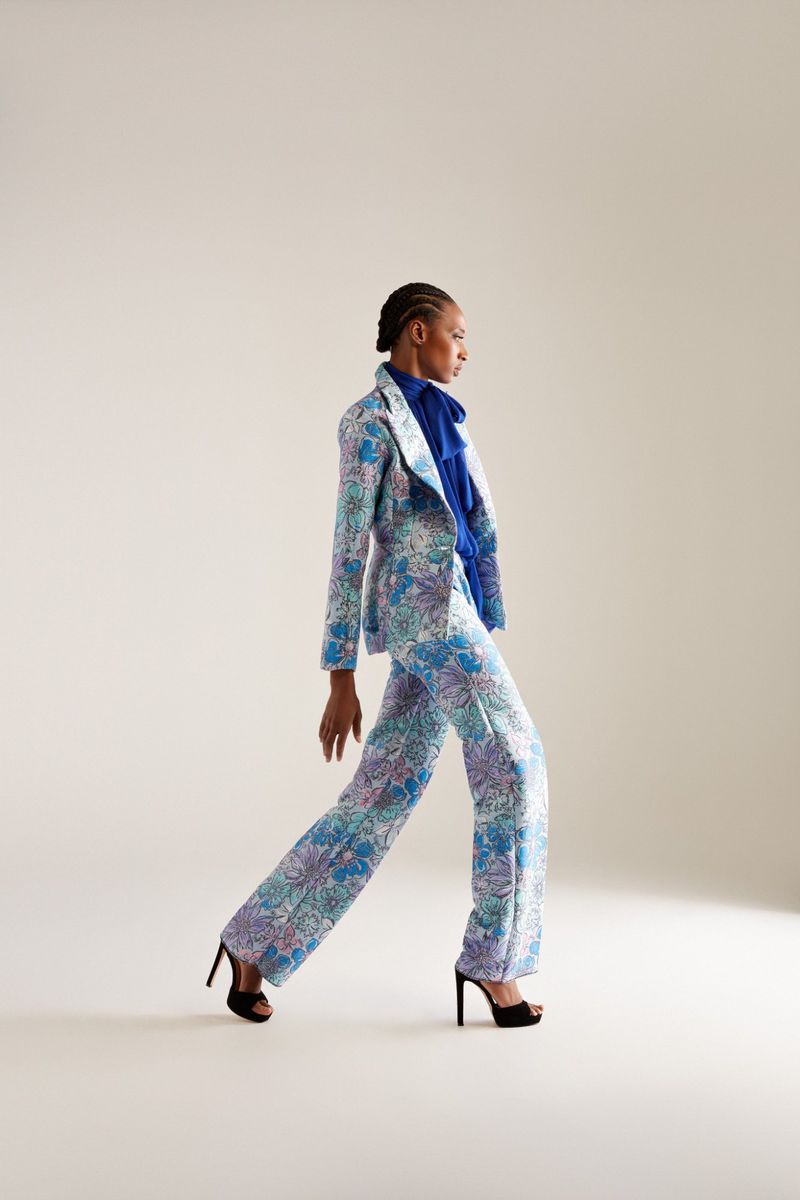 Woman in floral suit walks in a studio. She wears high heels, a blue top, and has braided hair, showcasing a fashion or editorial style.