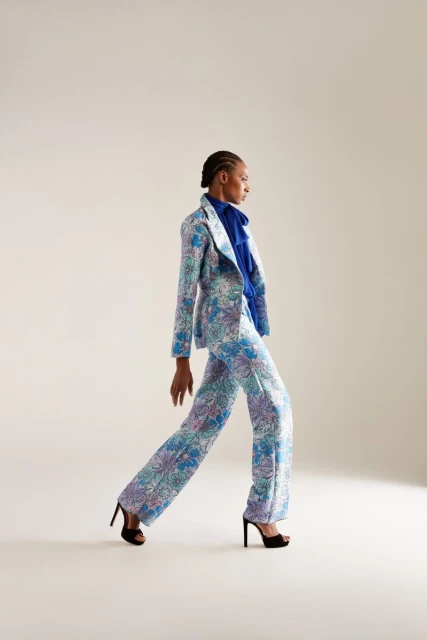 Woman in floral suit walks in a studio. She wears high heels, a blue top, and has braided hair, showcasing a fashion or editorial style.