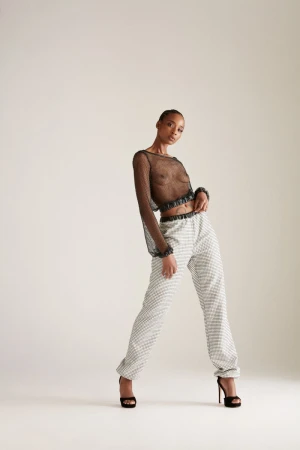 Model Goldie Carswell in a sheer black mesh long-sleeve crop top and patterned trousers with black heels against a white studio backdrop.