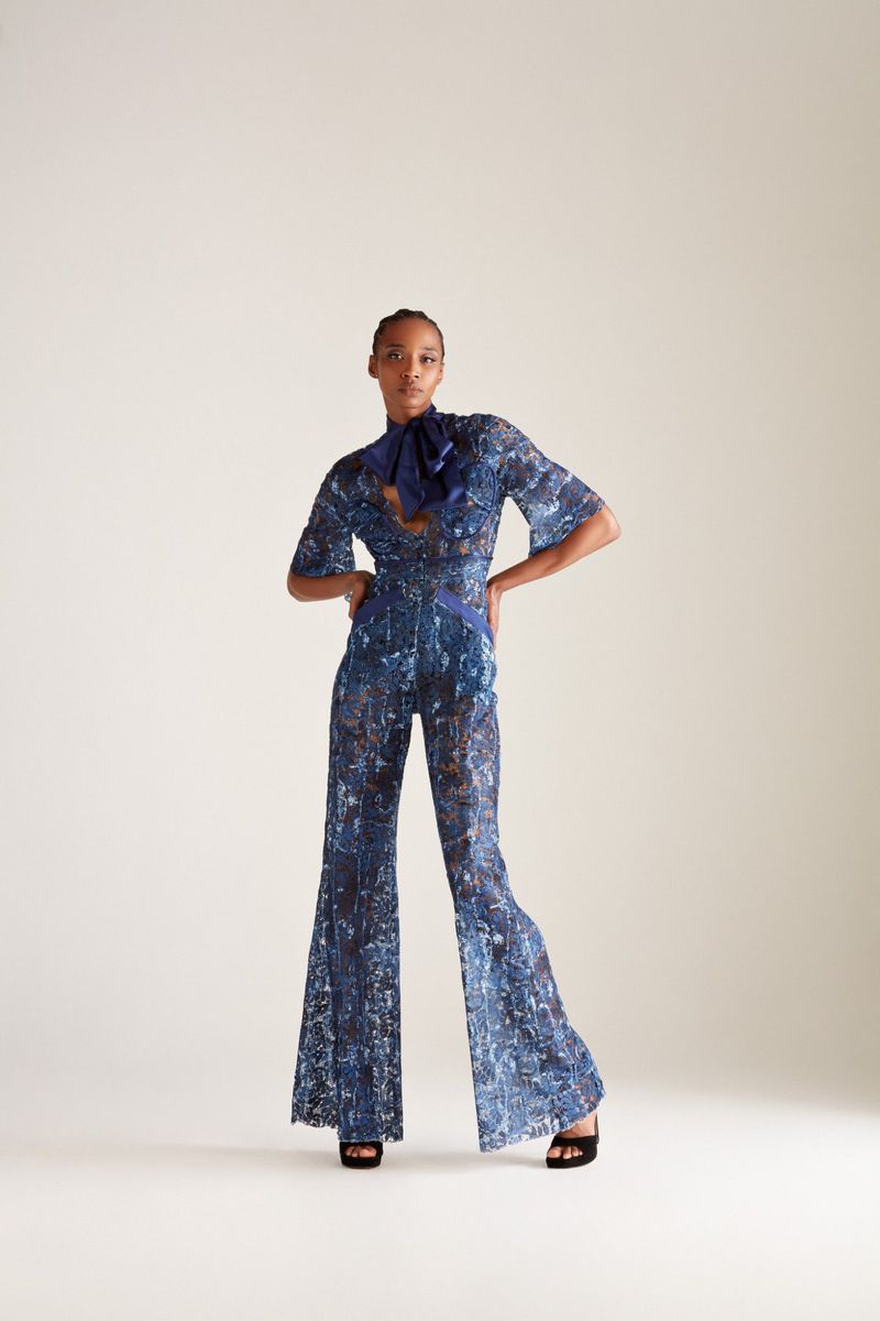 Full-length studio shot of an adult female fashion model wearing a blue floral-patterned jumpsuit with a pussy-bow collar and black high heels.