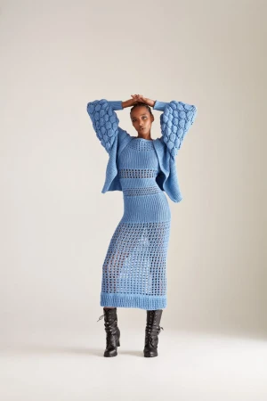 Model Goldie Carswell poses in a powder blue knit dress and matching textured cardigan with black boots against a white backdrop.