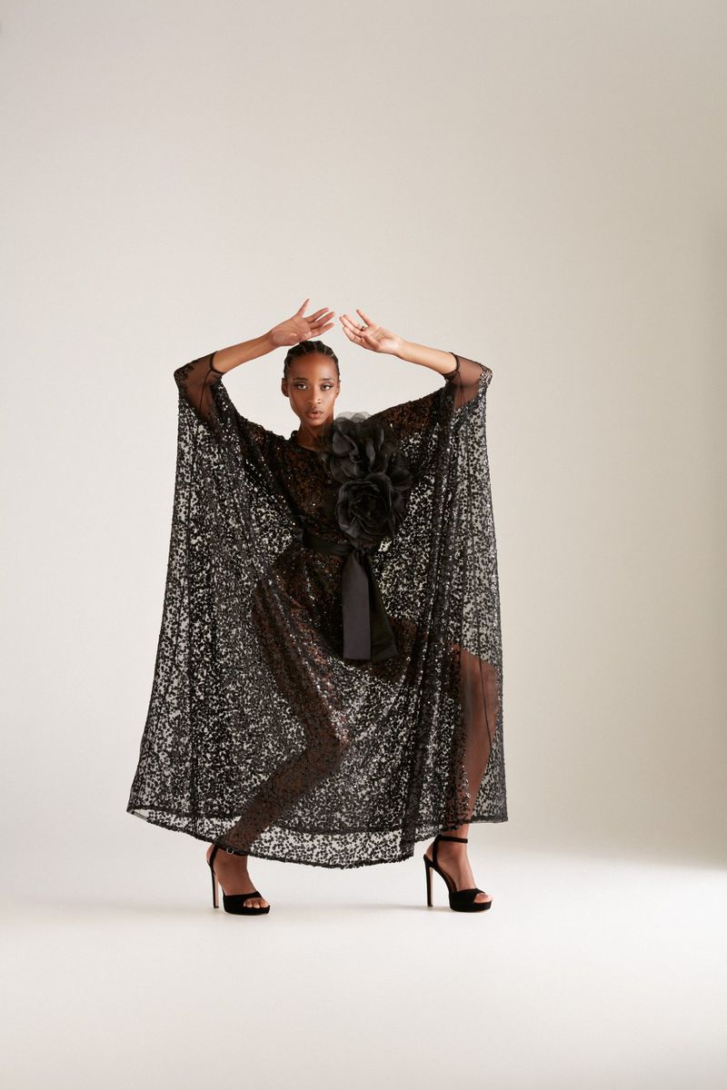 Elegant woman poses in a shimmering black gown with a floral accent and high heels against a neutral backdrop, fashion photoshoot.