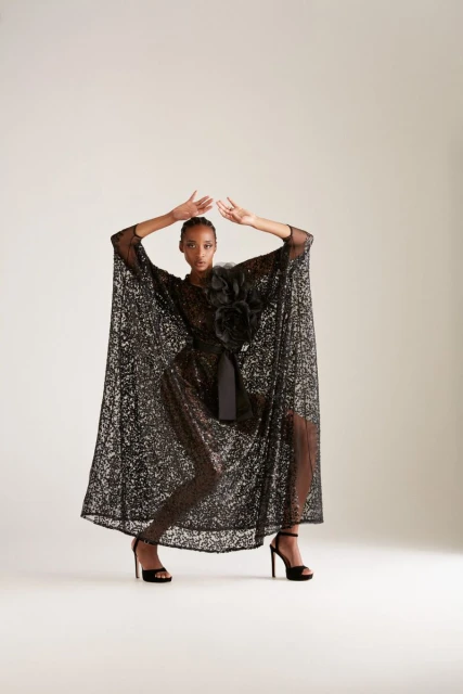 Elegant woman poses in a shimmering black gown with a floral accent and high heels against a neutral backdrop, fashion photoshoot.