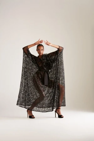 Elegant woman poses in a shimmering black gown with a floral accent and high heels against a neutral backdrop, fashion photoshoot.