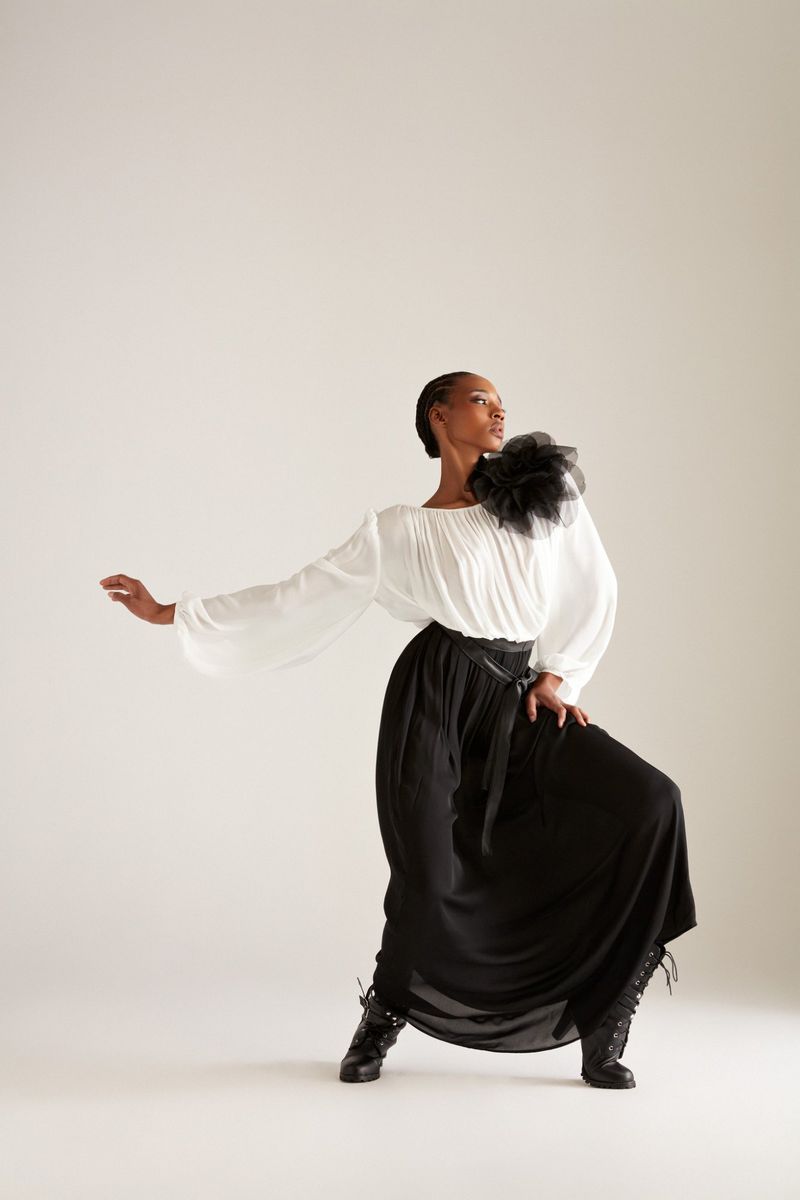 Model Goldie Carswell posing in a white blouse with a black shoulder rosette and long black skirt against a white studio backdrop.