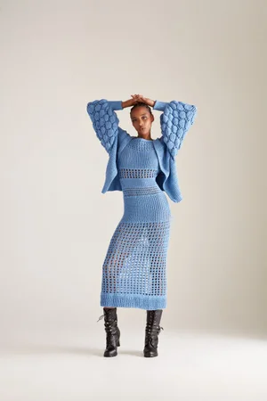 Model Goldie Carswell poses in a powder blue knit dress and matching textured cardigan with black boots against a white backdrop.