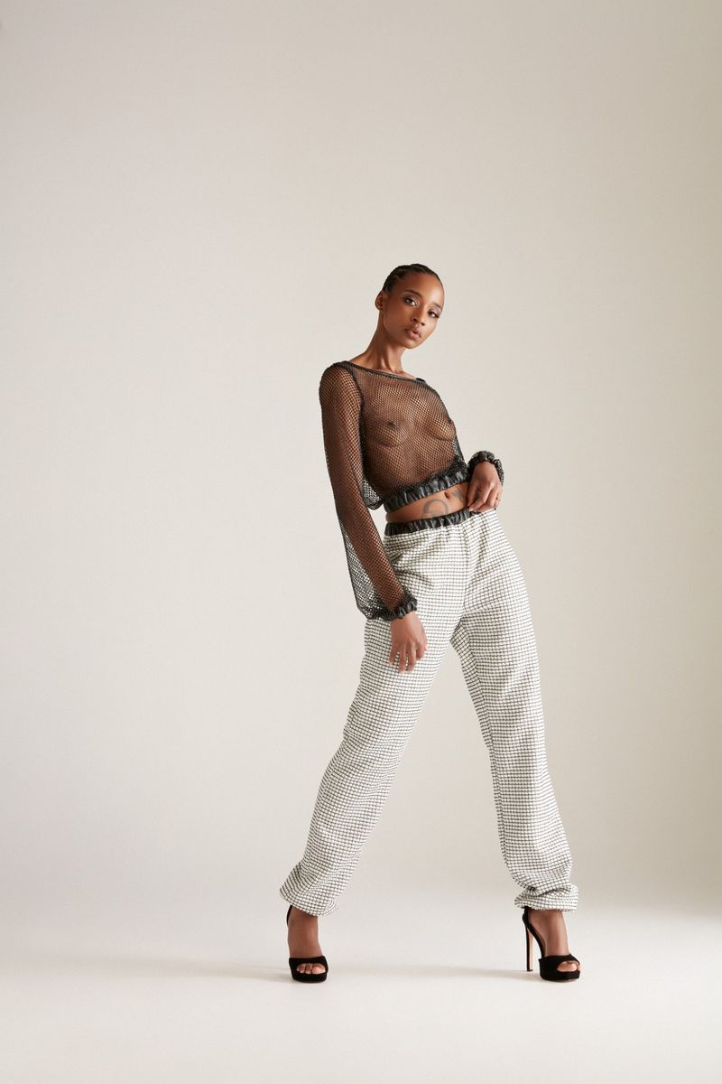 Model Goldie Carswell in a sheer black mesh long-sleeve crop top and patterned trousers with black heels against a white studio backdrop.