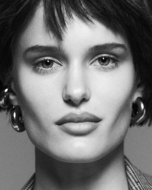 Close-up portrait of a young adult woman with short, dark hair and hoop earrings, wearing a jacket, in black and white.