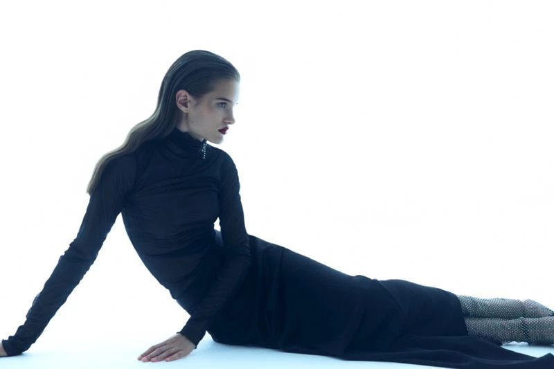 Fashion model in a black dress poses while lying down on a white backdrop. Long, wavy dark hair and red lipstick visible.