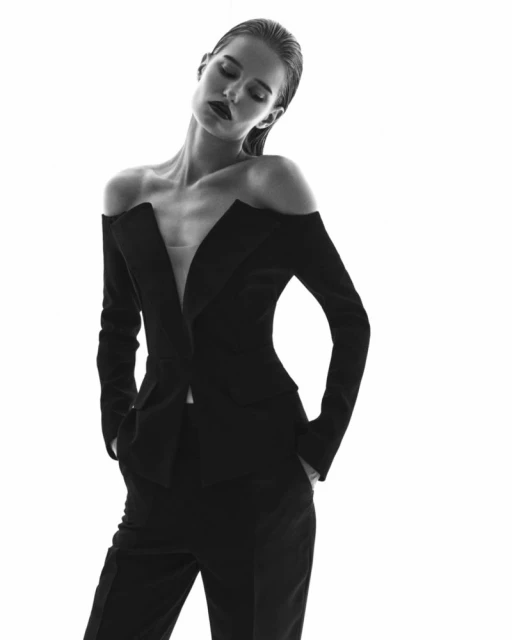 Monochrome portrait of a slender woman in an off-the-shoulder black blazer and pants suit against a bright white background.