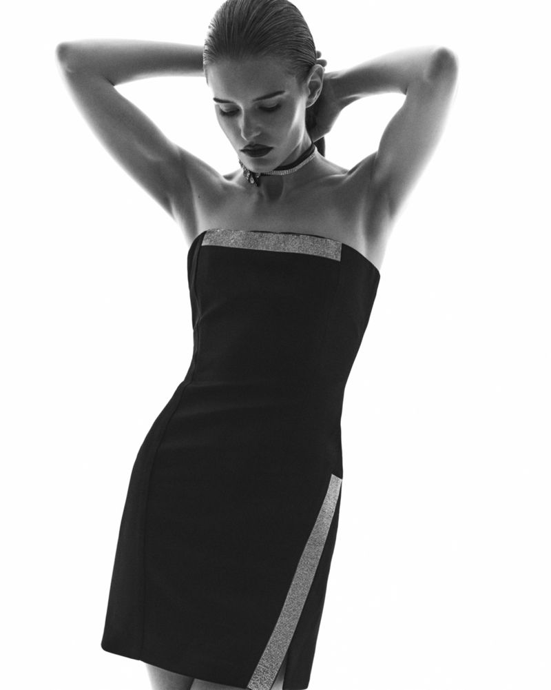Monochrome portrait of a slender woman in a black strapless dress, hands behind her head, against a stark white background in a photo studio.