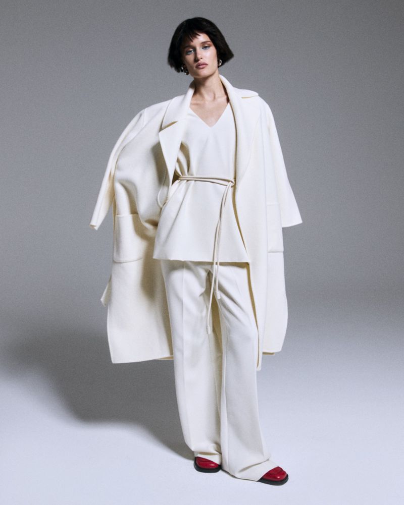 Fashion shot of a woman in a full-length off-white coat, blouse and wide-leg pants with red flats against a light gray background.