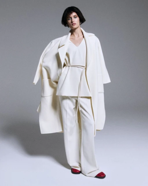 Fashion shot of a woman in a full-length off-white coat, blouse and wide-leg pants with red flats against a light gray background.