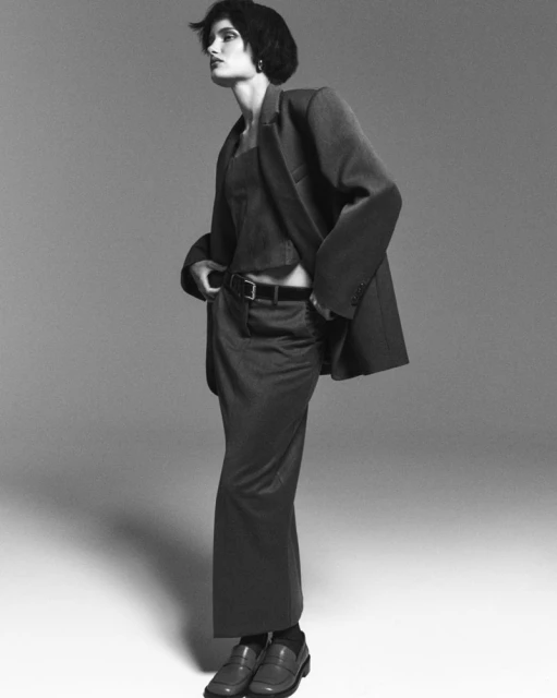 A woman in a dark skirt suit poses against a gray backdrop, looking off to the left. She is wearing leather loafers and dark socks.