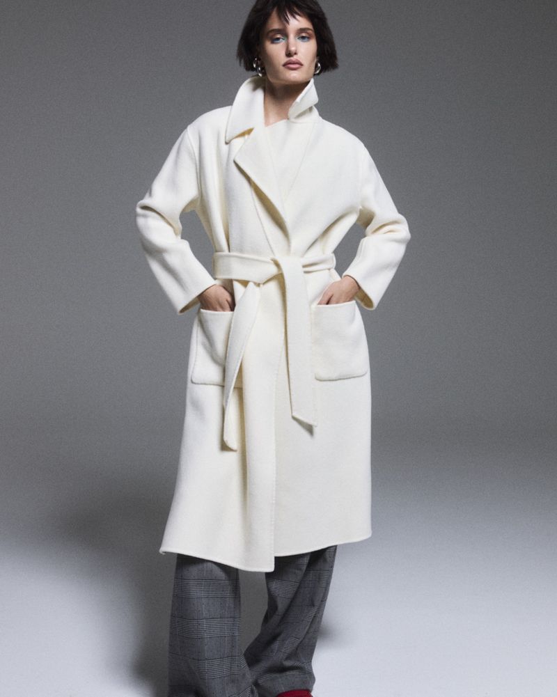 Model in an off-white belted wrap coat and grey plaid trousers posing against a grey studio backdrop.