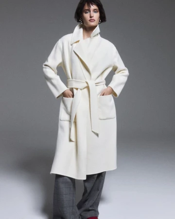 Model in an off-white belted wrap coat and grey plaid trousers posing against a grey studio backdrop.
