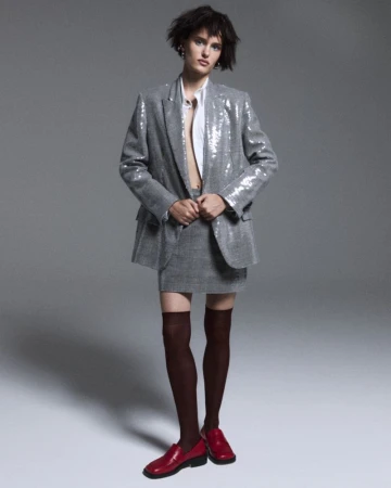 A young woman poses in a stylish grey sequined blazer and skirt, paired with knee-high socks and red loafers against a grey backdrop.