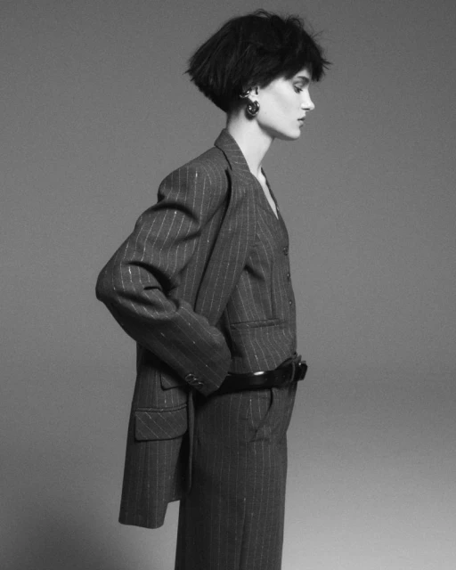 Monochrome studio shot of a young adult woman in a pinstripe suit with short dark hair, accessorized with large earrings and a belt.