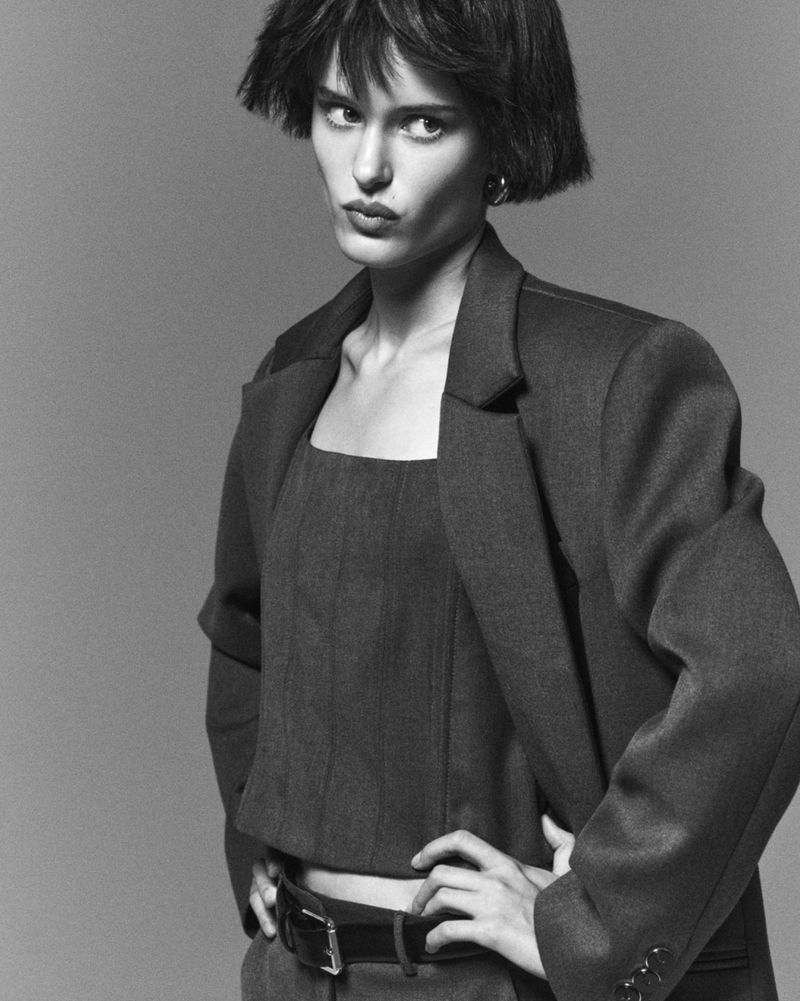 Monochrome portrait of a model with short hair wearing a jacket and top, hands on hips against a grey background in a studio shot.