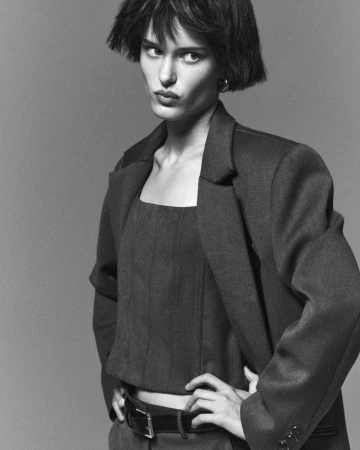 Monochrome portrait of a model with short hair wearing a jacket and top, hands on hips against a grey background in a studio shot.