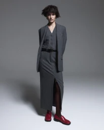 Model in a grey pinstripe suit vest, tailored blazer, and slit maxi skirt with red loafers against a grey studio backdrop.