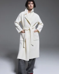 Model in an off-white belted wrap coat and grey plaid trousers posing against a grey studio backdrop.